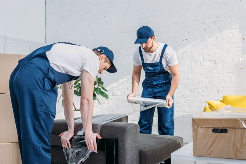 Two movers in blue overalls wrapping a piece of furniture in plastic wrap.