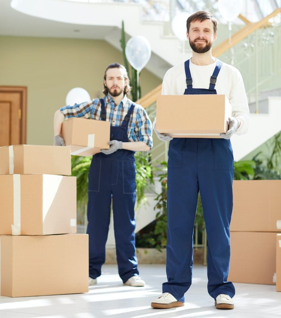 Two men with overalls holding boxes with 2 boxes next to them straight ahead at the camera.