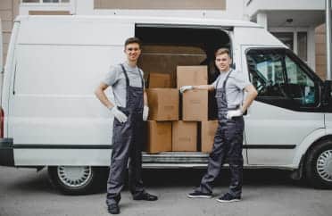 Two men posing in front of a moving truck filled with moving boxes inside the truck.
