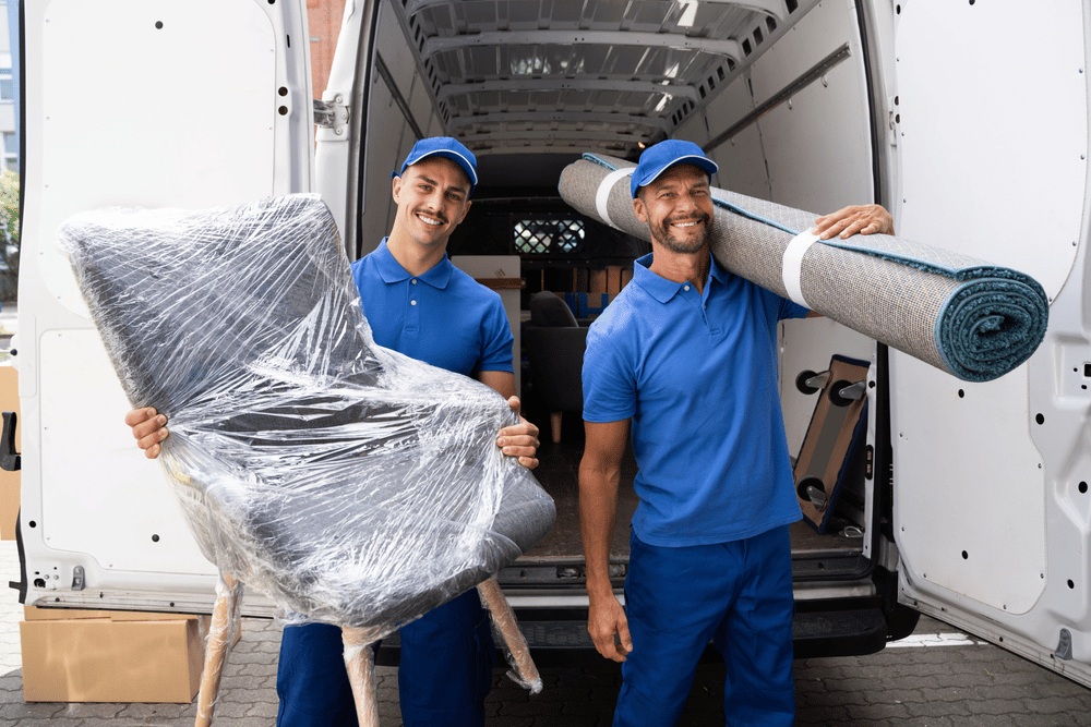 Two smiling men in blue moving a chair and a rolled up rug out of a moving van.